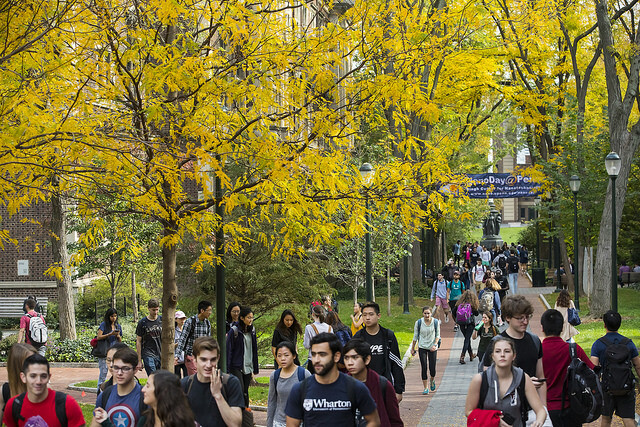 A group of students crowd Locust Walk between classes beneath colorful fall foliage